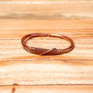 A handcrafted open-ended copper cuff bracelet resting on a wooden surface, featuring a smooth metallic band with a center section tightly wrapped in textured, twisted copper wire.