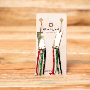 A pair of handcrafted drop earrings featuring a shiny, vertical rectangular golden brass bar from which four separate strands of small seed beads hang in red, white, green, and black representing the flag of Kenya.