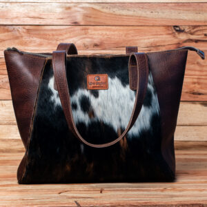 Handcrafted black and white cowhide tote with rich brown leather trim, shoulder straps, and zip-top closure.