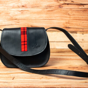 A black leather saddle-shaped crossbody bag featuring a vertical stripe of red and black plaid fabric down the center of its front flap and an adjustable leather shoulder strap.