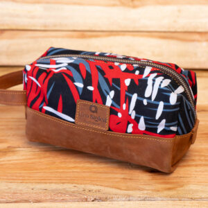 A rectangular toiletry pouch made from red, black, and white patterned African fabric with a brown leather base and side handle.