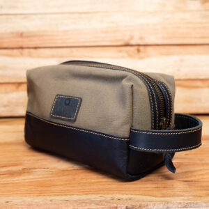 A rectangular canvas travel pouch in khaki green with a black leather base, yellow stitching, and a side handle.