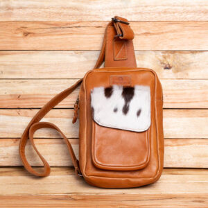 A tan leather crossbody sling bag featuring a spotted hair-on cowhide front pocket flap.