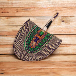 A colorful handwoven straw hand fan with a green, orange, and purple center, a patterned outer edge, and a leather-bound handle with a hanging loop.