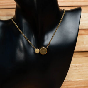 A gold-tone brass chain necklace featuring two circular disc pendants of different sizes, displayed on a black mannequin bust.