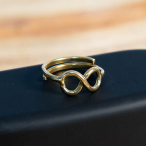 A gold-tone brass ring featuring an infinity symbol centerpiece, displayed on a black surface.